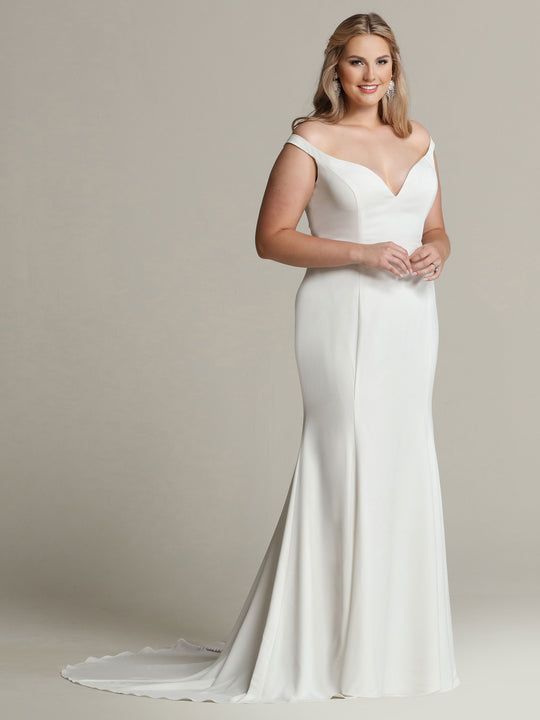 A woman in the Avery Austin Hazel ivory off-the-shoulder crepe wedding dress with a fitted bodice and slight train stands against a plain beige background, smiling gently.