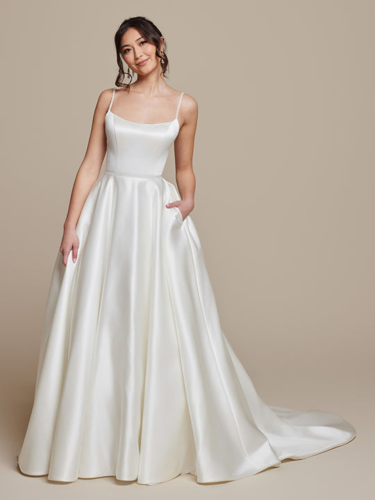 A woman in the averyaustin Millie wedding dress stands against a beige background, smiling as she holds the skirt of her modern ivory satin a-line gown with thin straps, a fitted bodice, and a long, flowing train.