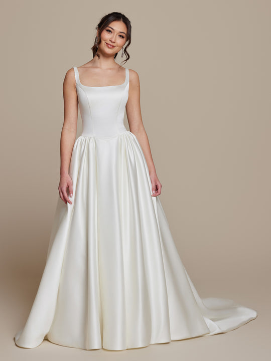 A woman wears the Tilly by averyaustin, an ivory satin wedding dress featuring a fitted bodice, square neckline, and full floor-length A-line skirt with a small train, set against a neutral background.