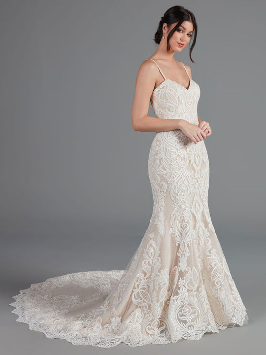 A woman models the Avery Austin Morgan wedding dress—a fitted, sleeveless lace gown with a mermaid silhouette, long train, and thin straps—against a gray background. Her elegant look is completed by an intricate pattern and low updo.