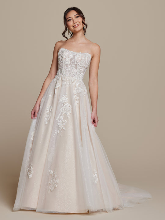 A woman smiles in the averyaustin Simone wedding dress, an ivory/nude glitter strapless corset A-line gown with beaded floral lace appliqué and subtle shimmer, elegantly displayed against a neutral beige background.