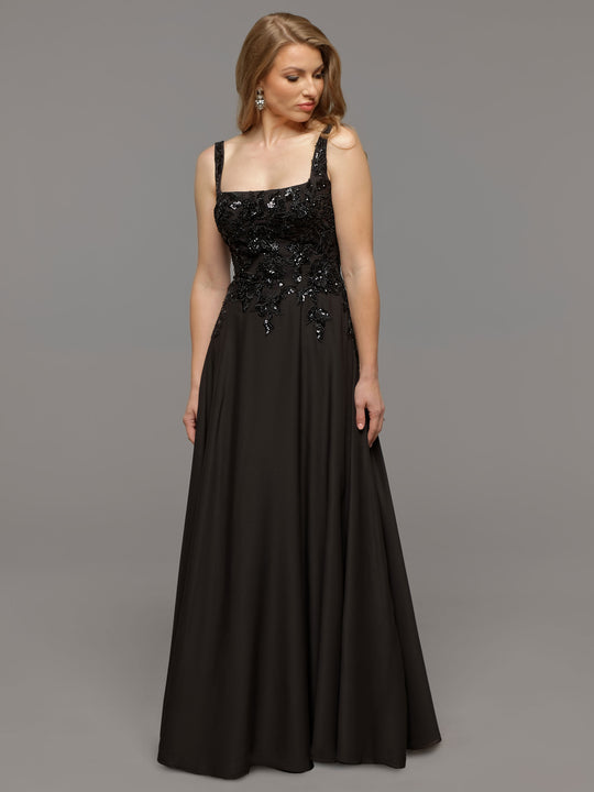 A woman models the averyaustin Scarlett gown in black, featuring a sleeveless design with a fitted embellished bodice and flowing A-line skirt, as she stands against a plain gray background looking down to her left.