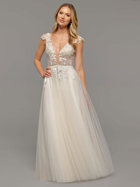 A woman wears the averyaustin Hana in Ivory/Ivory, an elegant floor-length lace wedding dress with a deep V-neck, sheer bodice, cap sleeves, and flowing tulle skirt, standing gracefully against a plain gray background.