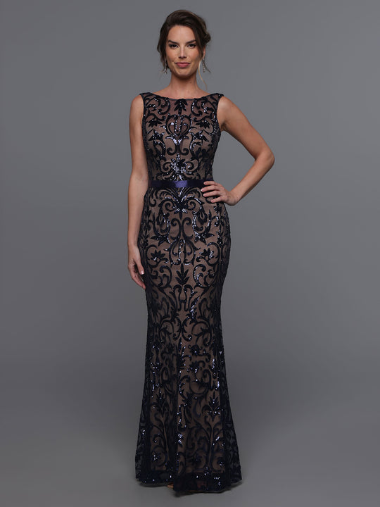 A woman stands against a gray background in the Avery Austin 80091 gown in Navy/Nude, featuring a floor-length sequin design with a sheer overlay, illusion neckline, and intricate shimmering patterns. She smiles softly with one hand on her hip.