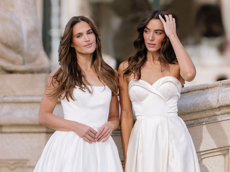 Two women in white Avery Austin bridal dresses standing outdoors with classical architecture in the background