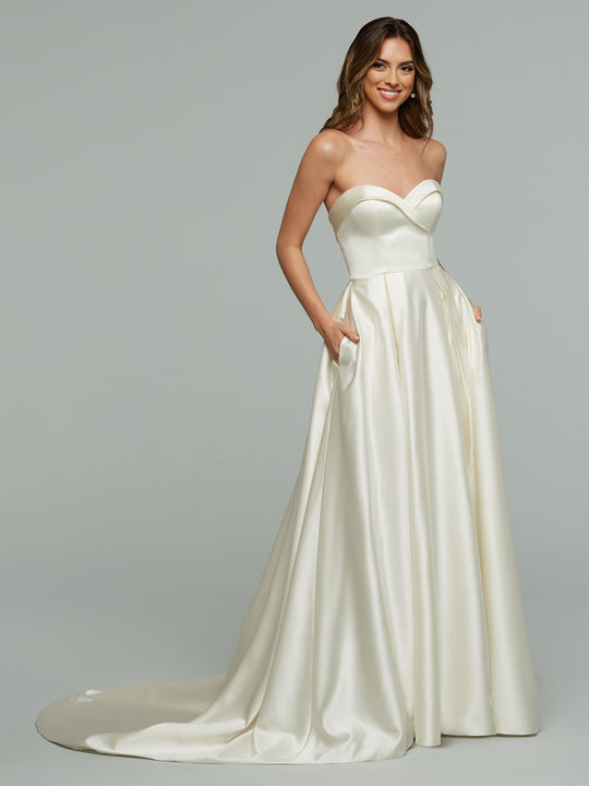 A woman smiles in the Avery Austin "Zara" gown, an ivory, strapless wedding dress with a sweetheart neckline, pockets, and pleated ball gown silhouette. The fitted bodice and full skirt with slight train are shown against a plain background.