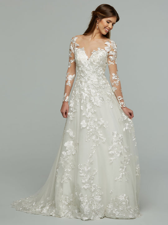 A woman smiles and looks to the side in the Avery Austin "Dylan" wedding dress, featuring ivory lace with floral appliqués and illusion sleeves, standing against a plain background.