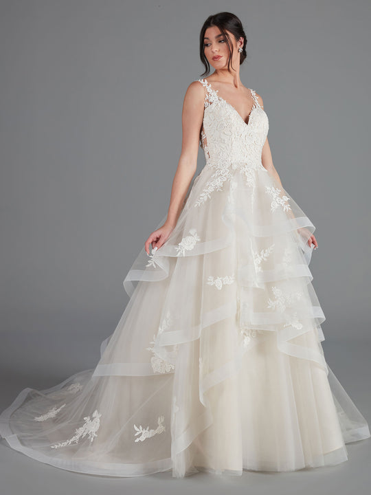 A woman models the Valencia by Avery Austin, an ivory/porcelain sleeveless V-neck lace tulle wedding dress with a layered skirt and floral appliqués, standing against a plain gray background.