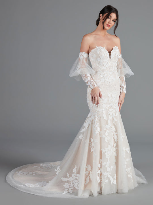 A woman models the Avery Austin "Kenya" wedding dress in Ivory/Blush, featuring off-the-shoulder lace, sheer long sleeves with floral patterns, a fit-and-flare mermaid silhouette, and a flowing train against a plain gray background.