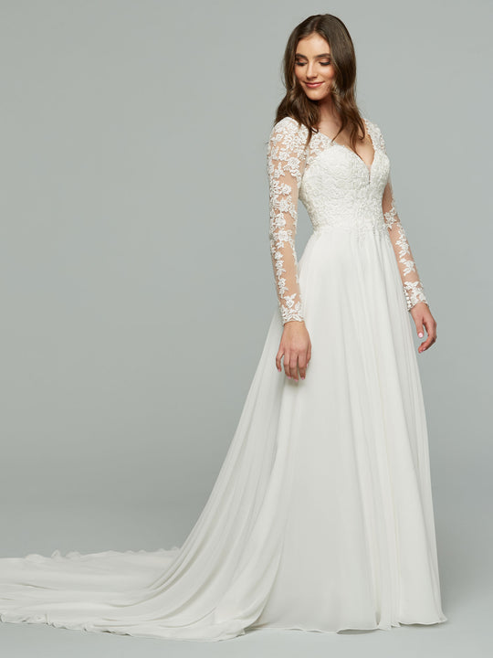 A woman in the Avery Austin Francesca wedding dress in ivory, featuring a delicate lace bodice and long lace sleeves, stands against a plain gray background, looking down and smiling softly.