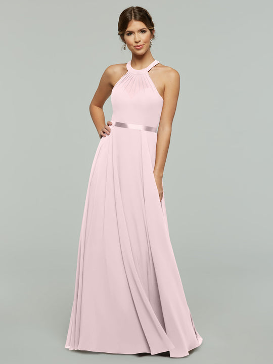 A woman models Avery Austin's 80038 dress, featuring an A-line chiffon design with a light pink pleated skirt and halter neckline, as she poses against a gray background with one hand on her hip and a gentle smile.