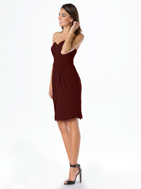 A woman smiles in Avery Austin's 80041 knee-length burgundy chiffon dress with a sweetheart neckline and black high-heeled sandals, posing against a white background as she touches her hair and looks to the side.