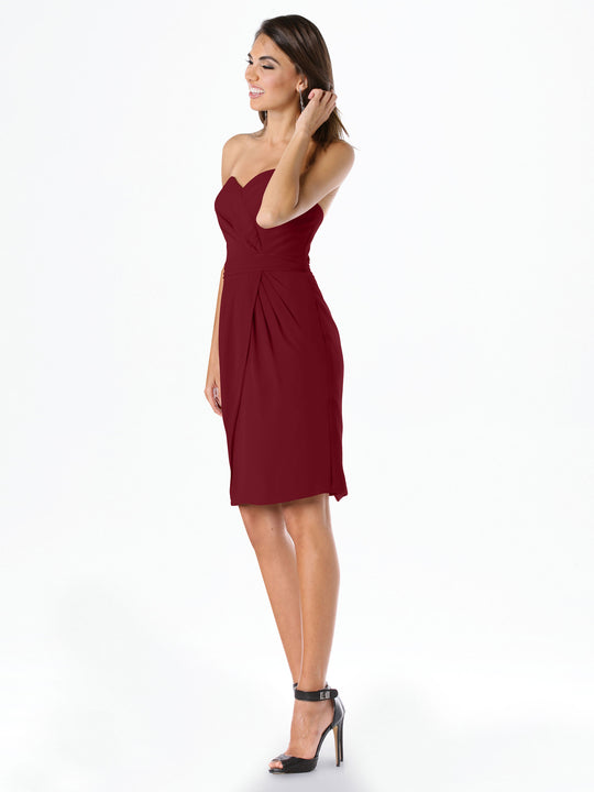 A woman poses against a plain white background, smiling in the Avery Austin 80041 strapless chiffon dress with pleats and a sweetheart neckline, paired with black high-heeled sandals as she touches her hair.