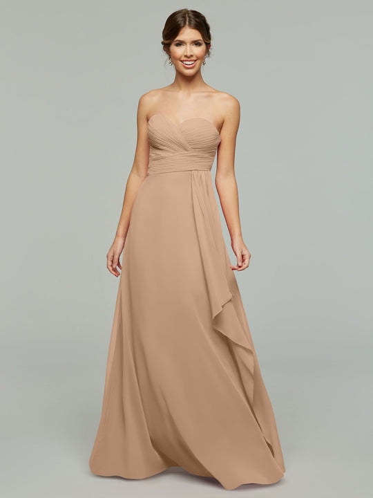 A woman smiles in front of a light gray background, wearing Avery Austin’s 80046: a strapless chiffon dress featuring a ruched bodice and flowing skirt, her hair styled in an elegant updo.