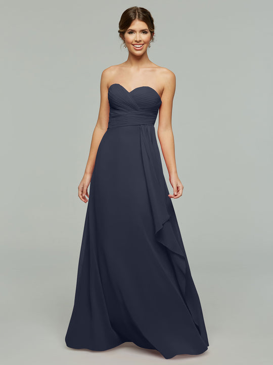 A woman smiles against a plain, light gray background, wearing Avery Austin's 80046—a timeless navy blue strapless chiffon dress with elegant draping and a flattering sweetheart neckline.