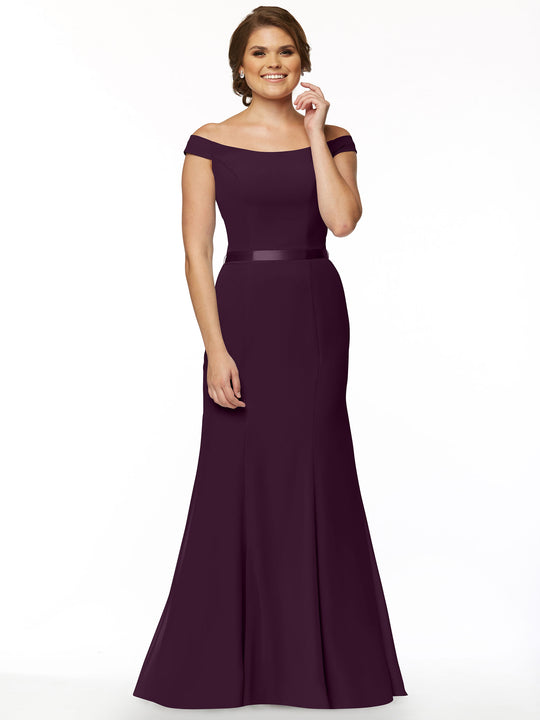 A woman smiles against a plain white background, wearing Avery Austin’s 80079 off-the-shoulder bridesmaid dress in aubergine with a fitted waist and elegant floor-length skirt.
