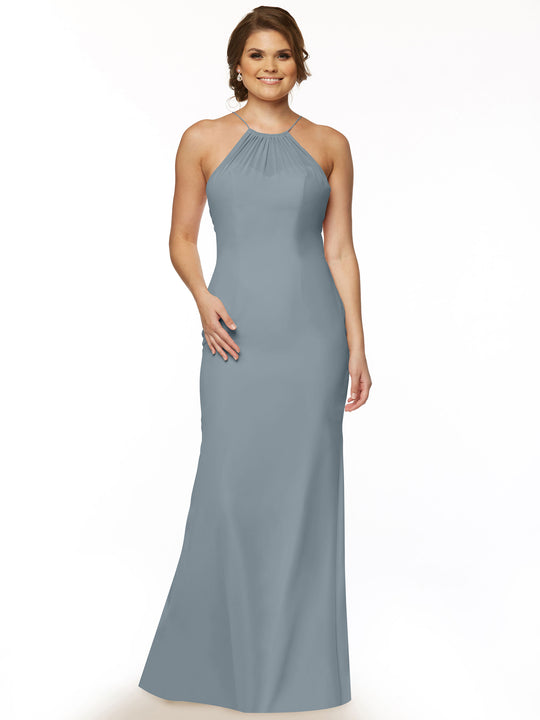 A woman stands smiling in Avery Austin’s 80083 gown—a sleeveless, floor-length chiffon dress in dusty blue with a halter neckline and chic keyhole back—set against a plain white background.