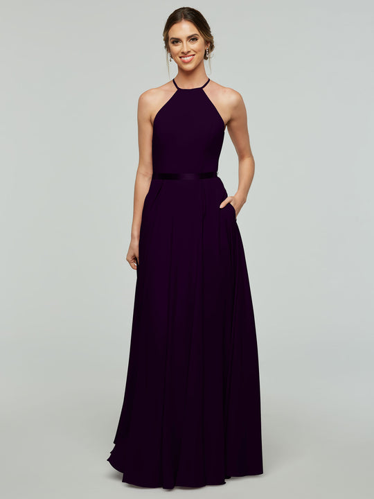 A woman stands against a light gray background, smiling with her hair pulled back, wearing the Avery Austin 80099—a floor-length, sleeveless dark purple A-line dress with a halter neckline and thin satin ribbon belt.