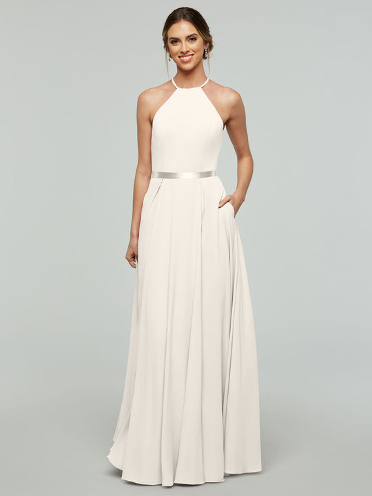 A woman models Avery Austin’s 80099—a sleeveless, white A-line dress with a halter neckline and satin ribbon belt. She smiles with light makeup, hair in a low bun, and poses with one hand in her pocket against a plain background.