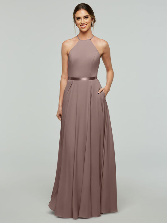 A woman wearing Avery Austin’s 80099, an A-line mauve gown with a halter neckline and satin ribbon belt, stands smiling with one hand in her pocket against a light gray background.