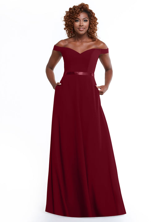 A woman with curly hair models Avery Austin's 80100: an off-the-shoulder, floor-length burgundy A-Line gown with a fitted bodice and satin waistband, standing against a plain white background.