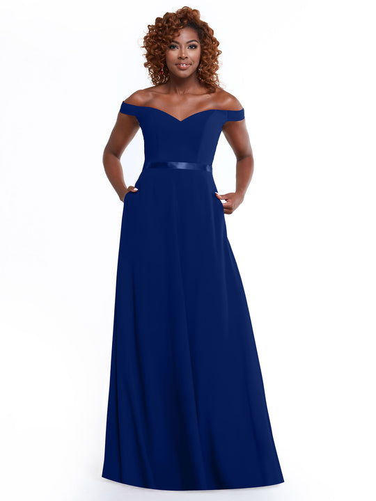 A woman with curly hair smiles in Avery Austin’s 80100 navy blue off-the-shoulder, floor-length A-line dress with a fitted bodice and pockets, standing against a plain white background.