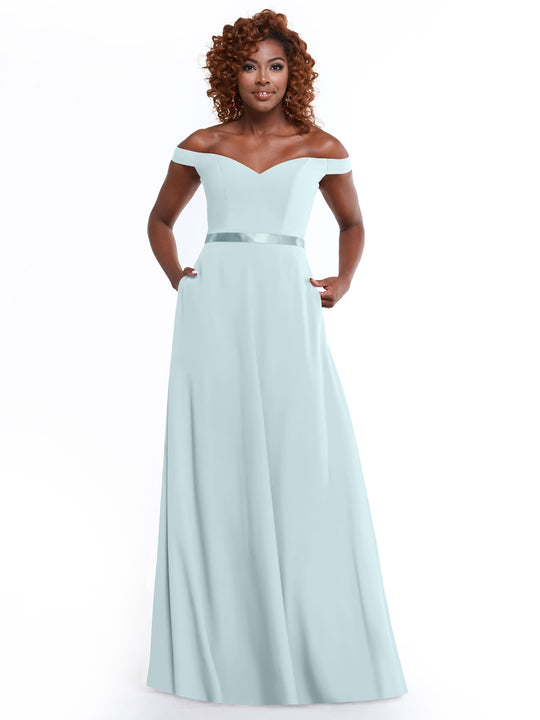 A woman smiles with her hands in the pockets of Avery Austin’s 80100 dress, a light blue, off-the-shoulder A-line style featuring a satin waistband, standing against a white background.
