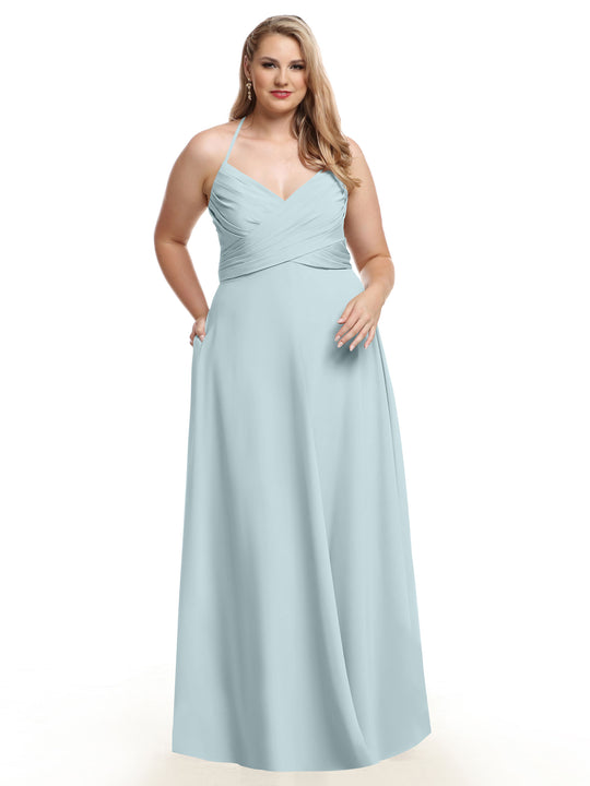 A woman with long, wavy blonde hair stands against a white background wearing the Avery Austin 80101 light blue A-line gown featuring a pleated bodice, V-neckline, and pockets.