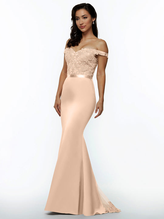 A woman models Avery Austin's 80104 blush bridesmaid dress, floor-length and off-the-shoulder with a lace bodice and satin waistband, standing before a plain white background.