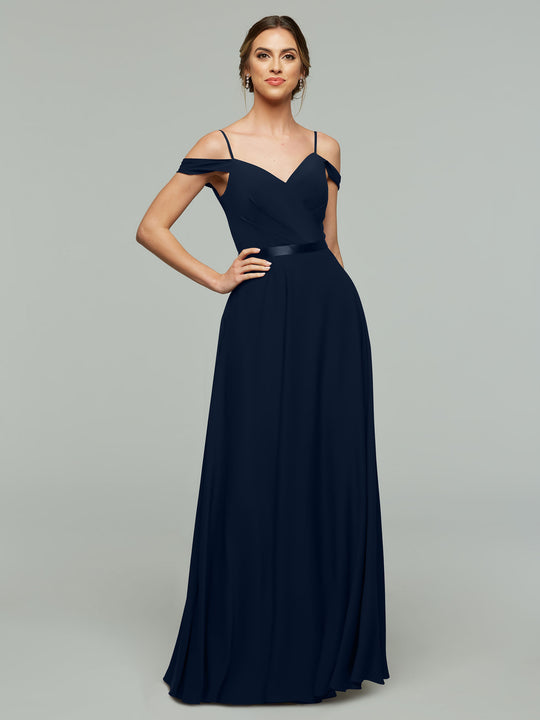 A woman models Avery Austin's floor-length, navy blue off-the-shoulder bridesmaid dress (style 80106) with thin straps and a fitted bodice against a plain light gray background.