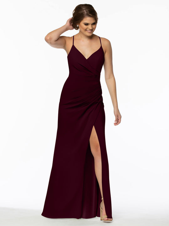 A woman models the Avery Austin 80114 bridesmaid dress—a sleeveless, deep burgundy v-neck gown with thin straps, a ruched waist, and a high slit. She pairs it with nude heels against a white background.