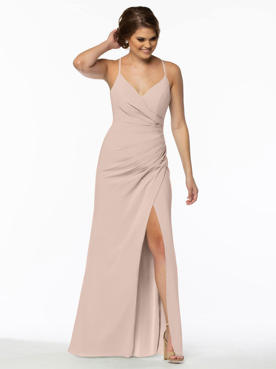 A woman in the Avery Austin 80114 rose pink chiffon bridesmaid dress with a V-neckline, ruched waist, and thigh-high slit stands smiling, her left hand touching her hair. She wears heels and poses against a plain white background.