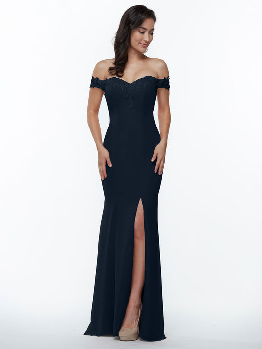 A woman in the Avery Austin 80121 midnight dress with off-the-shoulder lace details and a thigh-high slit stands against a white background, smiling as she looks down with her hands by her sides.
