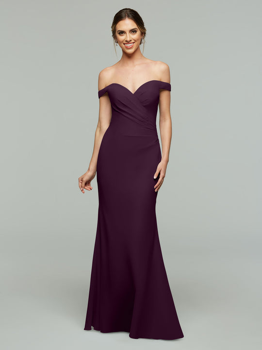 A woman stands smiling against a light gray background, wearing Avery Austin’s 80122 floor-length, off-the-shoulder bridesmaid dress in aubergine with a fitted bodice.