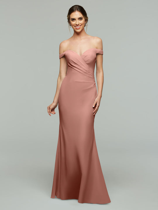 A woman wearing Avery Austin’s 80122 blush pink off-the-shoulder bridesmaid dress with a fitted bodice and flowing skirt smiles against a plain light gray background.