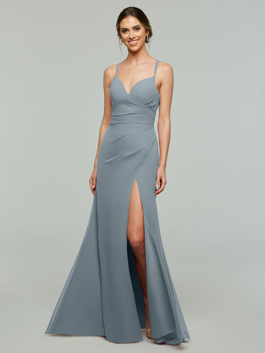 A woman is smiling and standing against a light gray background, wearing the Avery Austin 80130 bridesmaid dress in Dusty Blue—a sleeveless, floor-length chiffon gown with spaghetti straps, a ruched bodice, v-neckline, and high side slit.