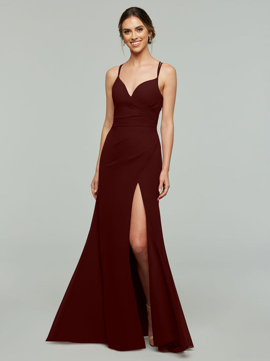 A woman smiles against a light gray background wearing Avery Austin style 80130, a merlot sleeveless floor-length sheath bridesmaid dress with a V-neck, thin straps, and high slit.