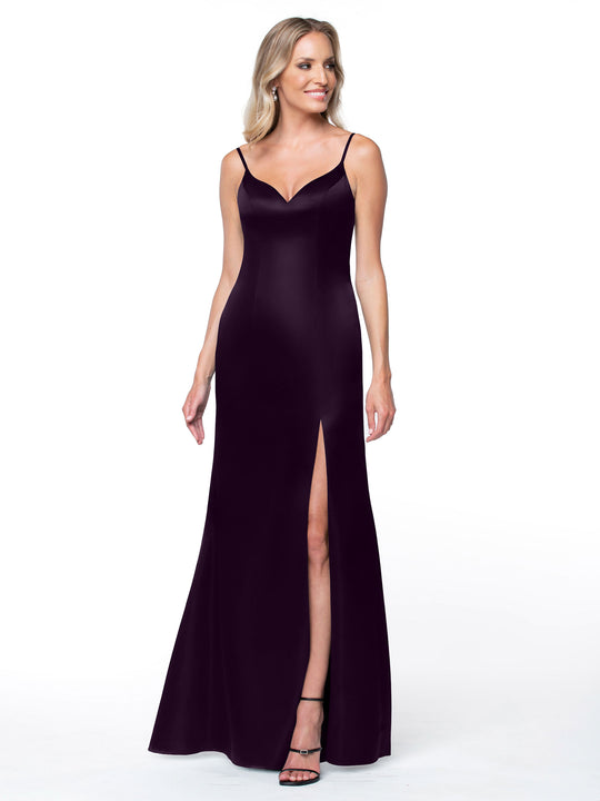 A blonde woman stands smiling on a white background, wearing Avery Austin style 80132—a long, sleeveless deep purple dress with thin straps, a thigh-high slit, and black high-heeled sandals.