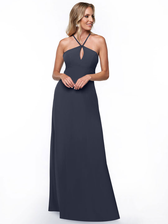 A woman with light hair smiles softly, wearing Avery Austin style 80137—a floor-length, dark blue halter dress with a keyhole neckline—against a plain white background. Her hands are clasped, highlighting the dress’s minimalistic elegance.