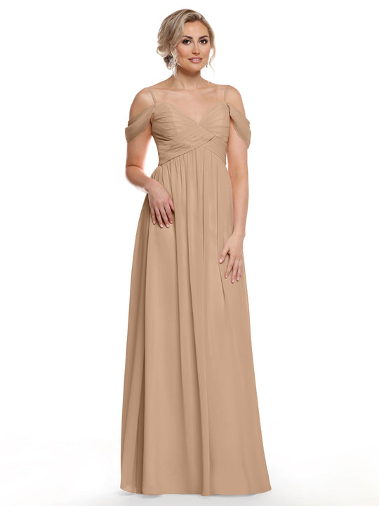 A woman models Avery Austin style 80142, a long buff chiffon cold shoulder dress with a pleated bodice and sheer straps. Her blonde hair is styled up and she pairs the look with statement earrings against a white backdrop.