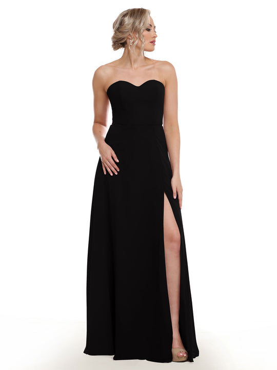 A woman stands against a plain white background wearing Avery Austin’s 80143: a black, floor-length strapless sweetheart gown with a sash back and side slit.