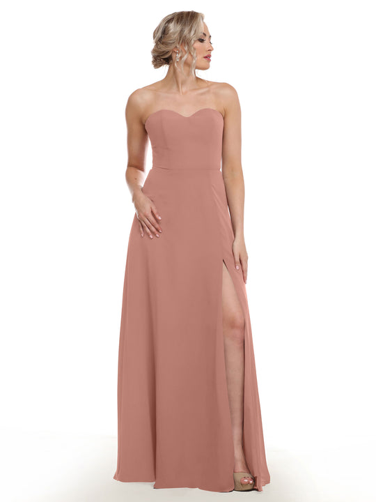 A woman wearing Avery Austin’s 80143, a blush pink strapless sweetheart dress with a sash back, floor-length silhouette, and side slit, stands looking to the side against a white background.