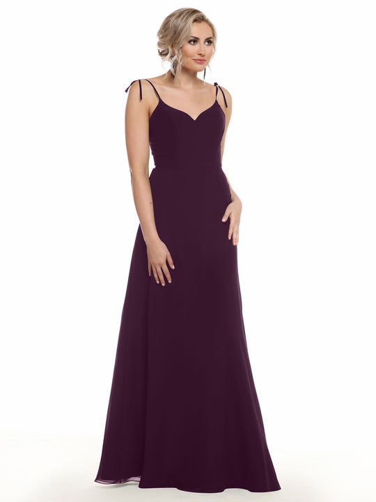 A woman wears the Avery Austin 80144, a floor-length, sleeveless deep purple V-neck chiffon dress with thin straps. She stands against a white background, with blonde waves and one hand at her side while the other touches her hip.