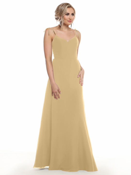 A woman models Avery Austin’s 80144, a long light yellow chiffon dress with spaghetti straps, standing against a white background, gazing to the side with one hand by her side and the other touching her skirt.