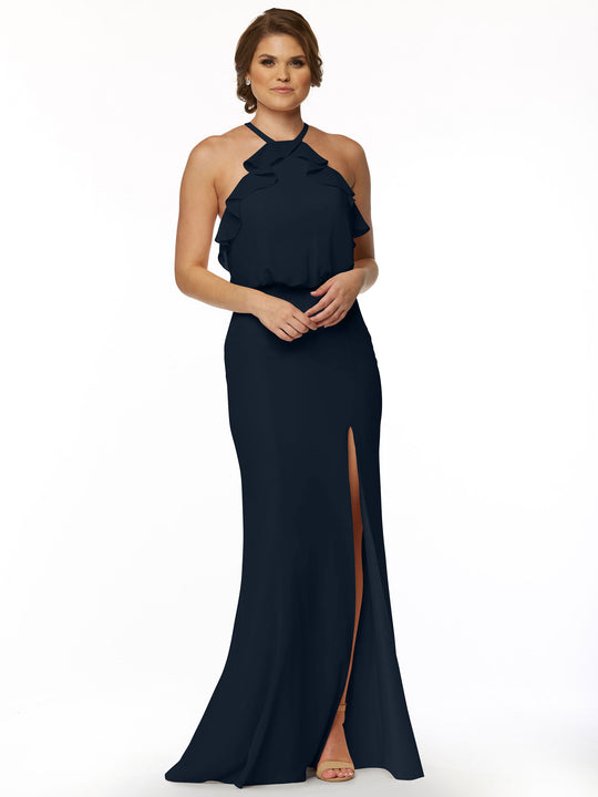 A woman with short hair wears Avery Austin’s 80147, a fitted chiffon gown featuring a ruffled halter top, floor-length black skirt with a front slit, and nude heels against a plain white background.