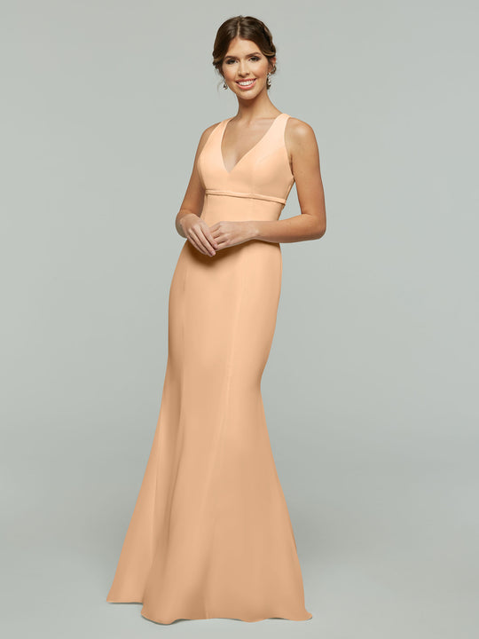 A woman wearing Avery Austin’s 80155 sleeveless blush fit and flare chiffon dress with an open keyhole back stands against a light gray background, smiling with her hands gently clasped in front of her.