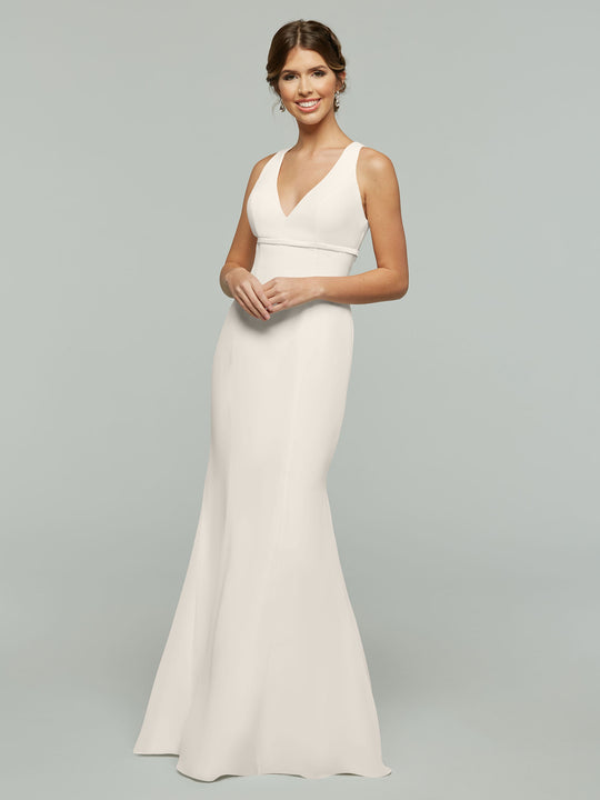 A woman smiles against a plain light gray background, wearing Avery Austin’s 80155—a sleeveless white fit and flare gown with a V-neckline, fitted waist, and satin ribbon accent.