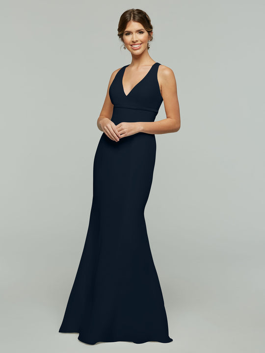 A woman smiles and clasps her hands in front of her while modeling Avery Austin style 80155, a sleeveless floor-length navy chiffon dress with a V-neckline and open keyhole back, against a plain light gray background.