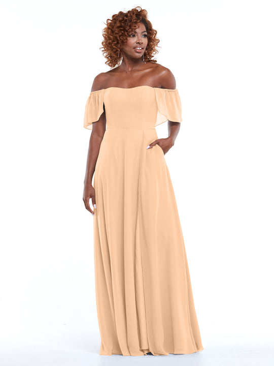 A woman with curly hair stands against a white background, gazing to the side in the Avery Austin 80168 blush off-the-shoulder chiffon A-line dress with pockets, one hand casually tucked into her pocket.