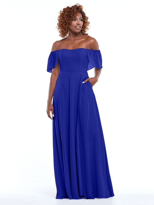 A woman with curly hair stands against a white background, gazing to the side in Avery Austin’s cobalt chiffon off-the-shoulder A-Line dress (style 80168) with flutter sleeves and pockets, one hand tucked casually in her pocket.